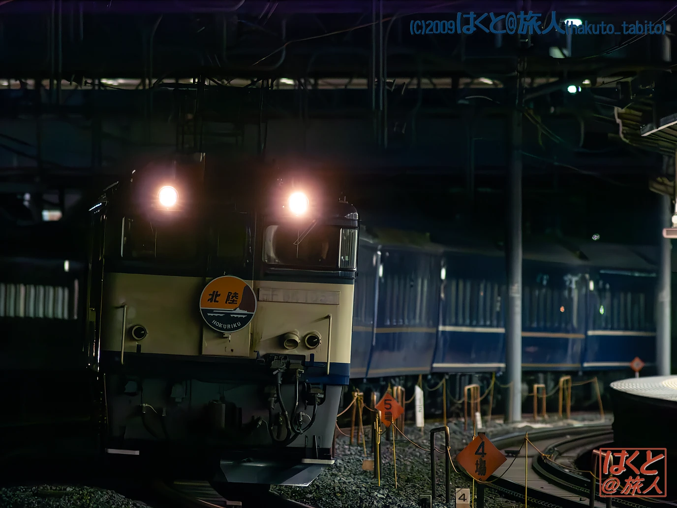 Front view of a Hokuriku railway locomotive at night on a yard track, headlights on, blue passenger cars in the background