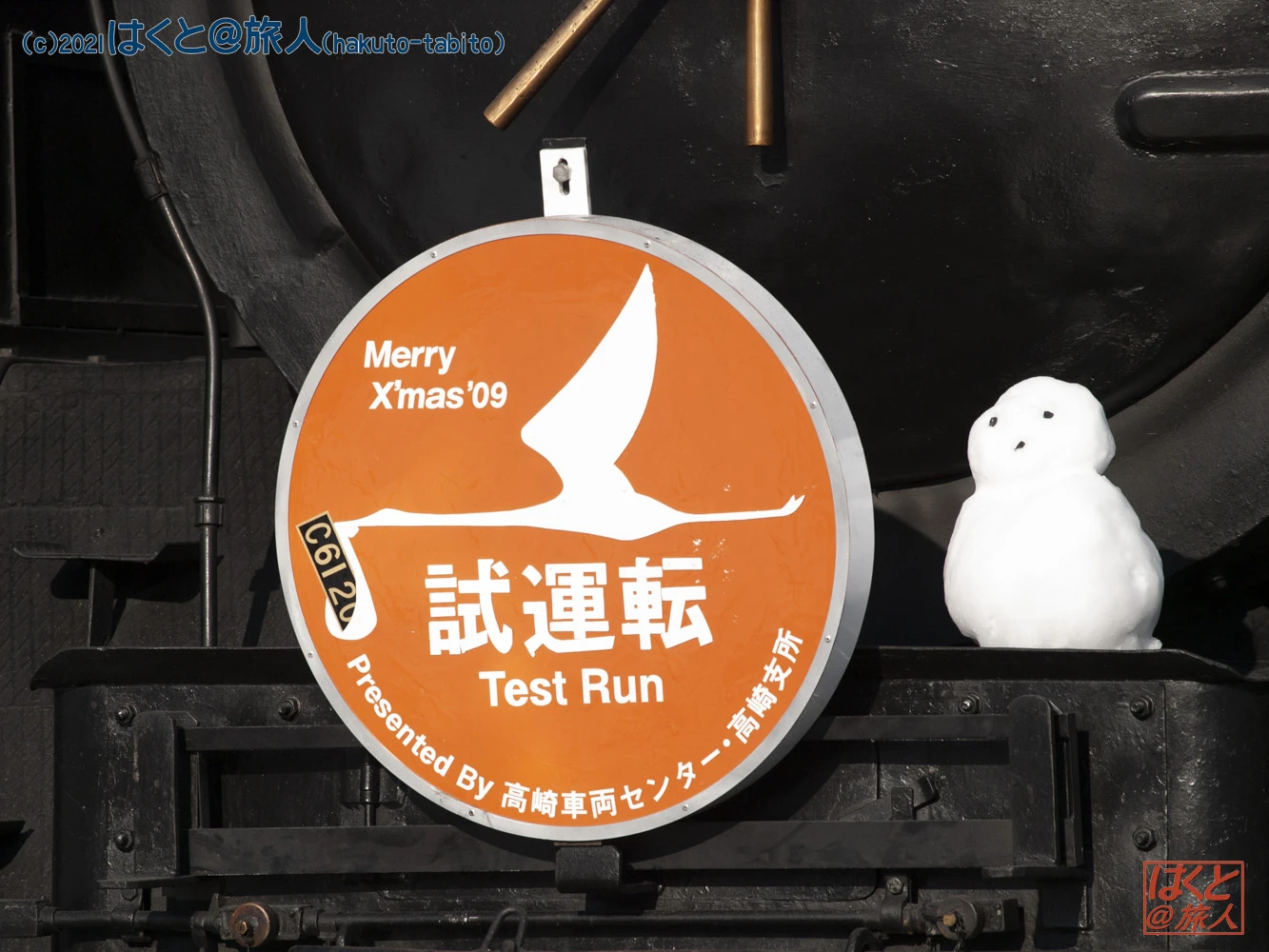 Orange circular sign on a train reading 'Merry Xmas ’09' and 'Test Run' with a white bird silhouette; a small snowman sits to the right.