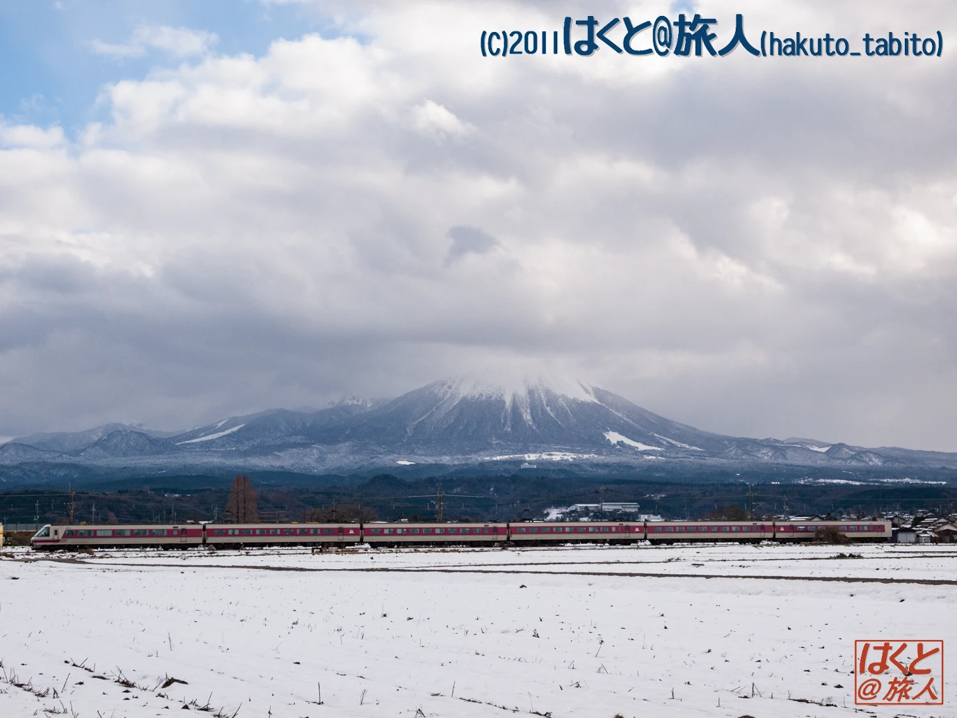 Long red-and-silver passenger train trailing across a snow-covered plain with a snow-dusted mountain range in the distance under a cloudy sky