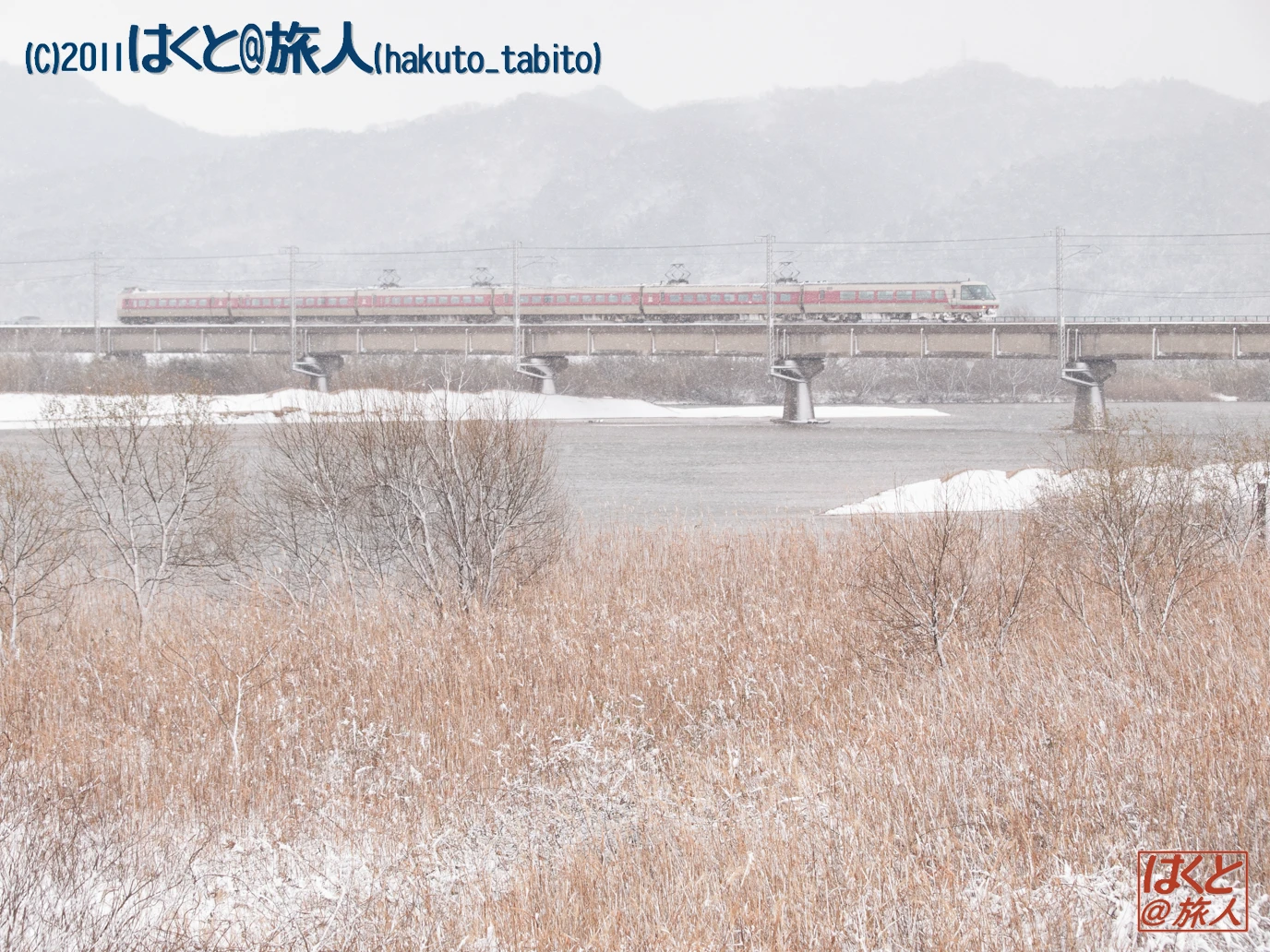 Passenger train crossing a snow-covered river on a bridge, with bare reeds in the foreground and distant mountains in the background.
