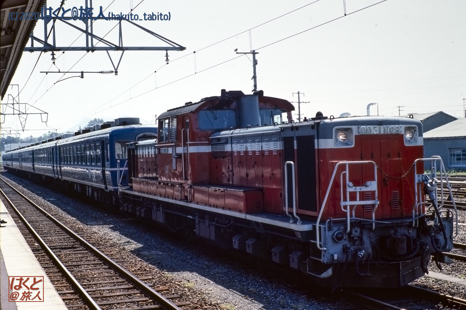 Red diesel locomotive pulling blue passenger cars at a sunny train station, with overhead wires and a platform on the left.