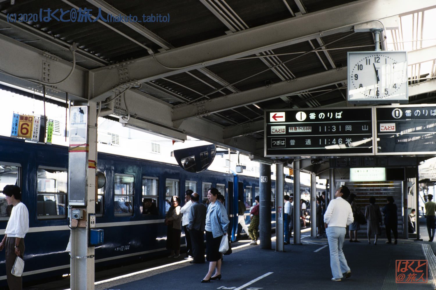 People wait on a train platform beside a blue commuter train; a large clock and timetable board hang overhead.