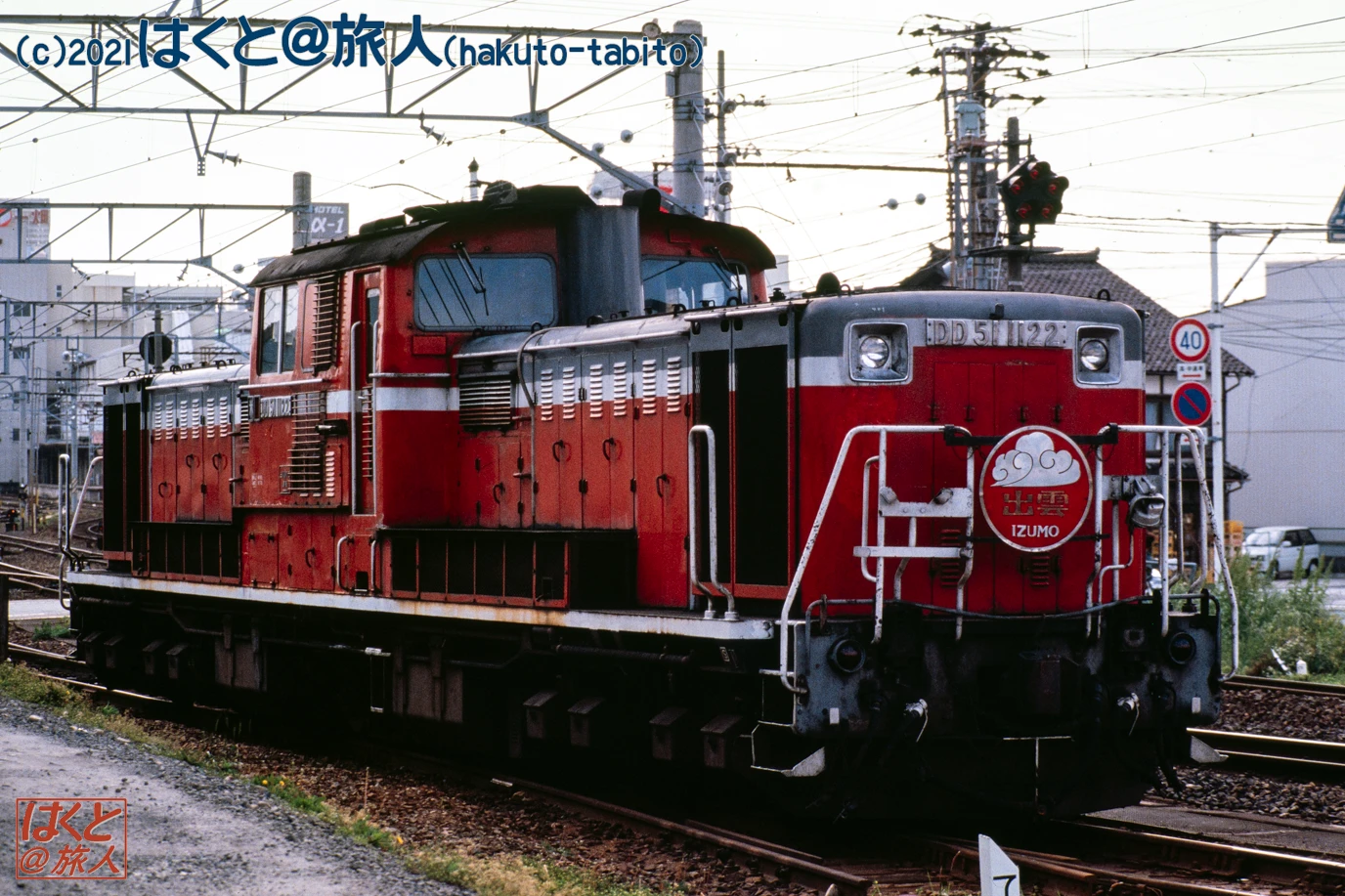 Red diesel locomotive on a railway track, facing the camera, with the Izumo emblem on the nose amid urban surroundings and overhead wires.