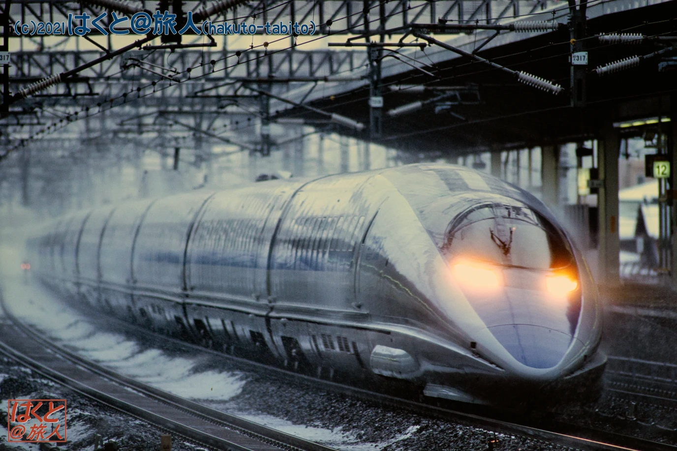 Front view of a silver high-speed train speeding along wet tracks at a station, headlights glowing, overhead wires overhead.