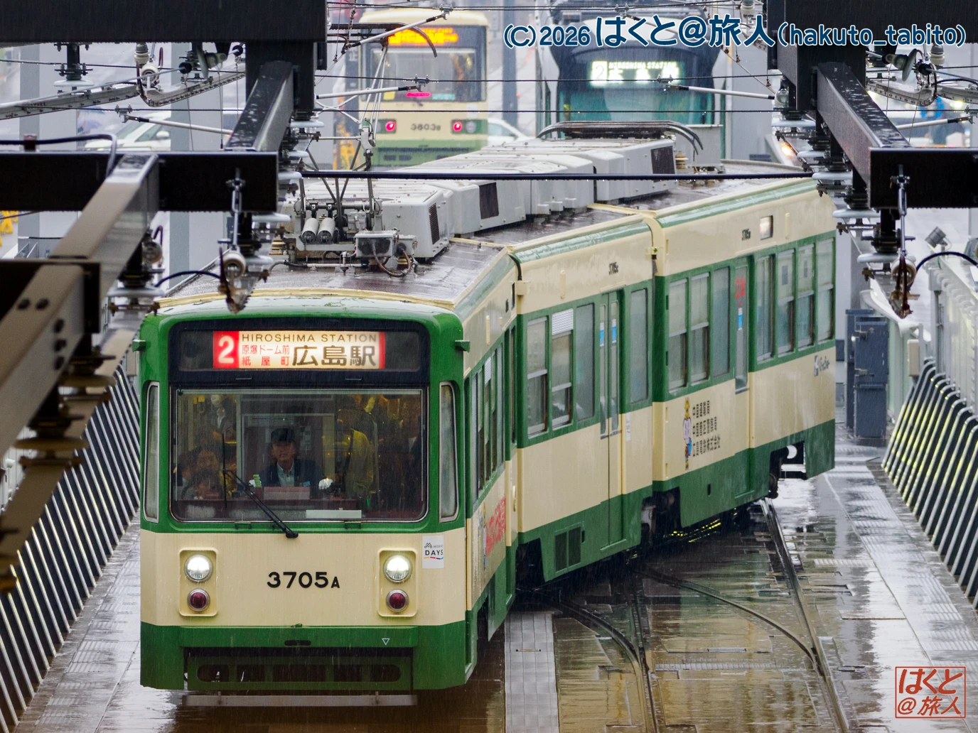 Green and cream tram at a rainy station platform, front view with an operator visible inside; destination sign reads Hiroshima Station.