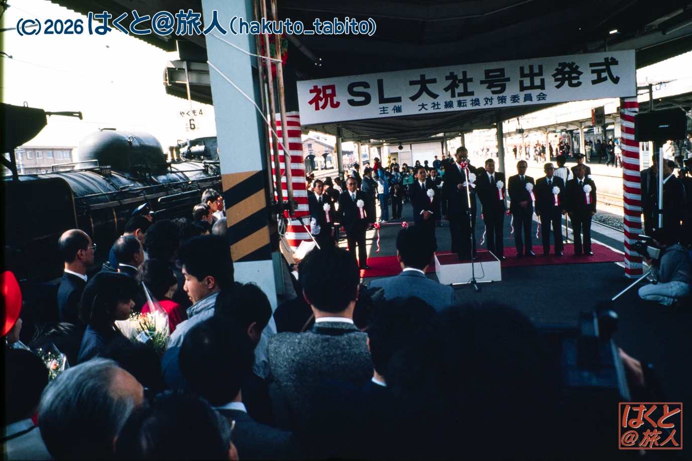Officials in suits on a stage at a train platform during a departure ceremony, with a steam locomotive on the left and a watching crowd.