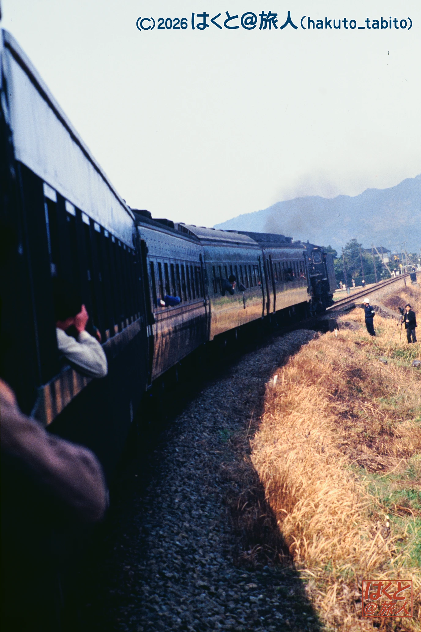 Long blue passenger train on tracks beside dry grass, with distant mountains and a person leaning from a window on the left.