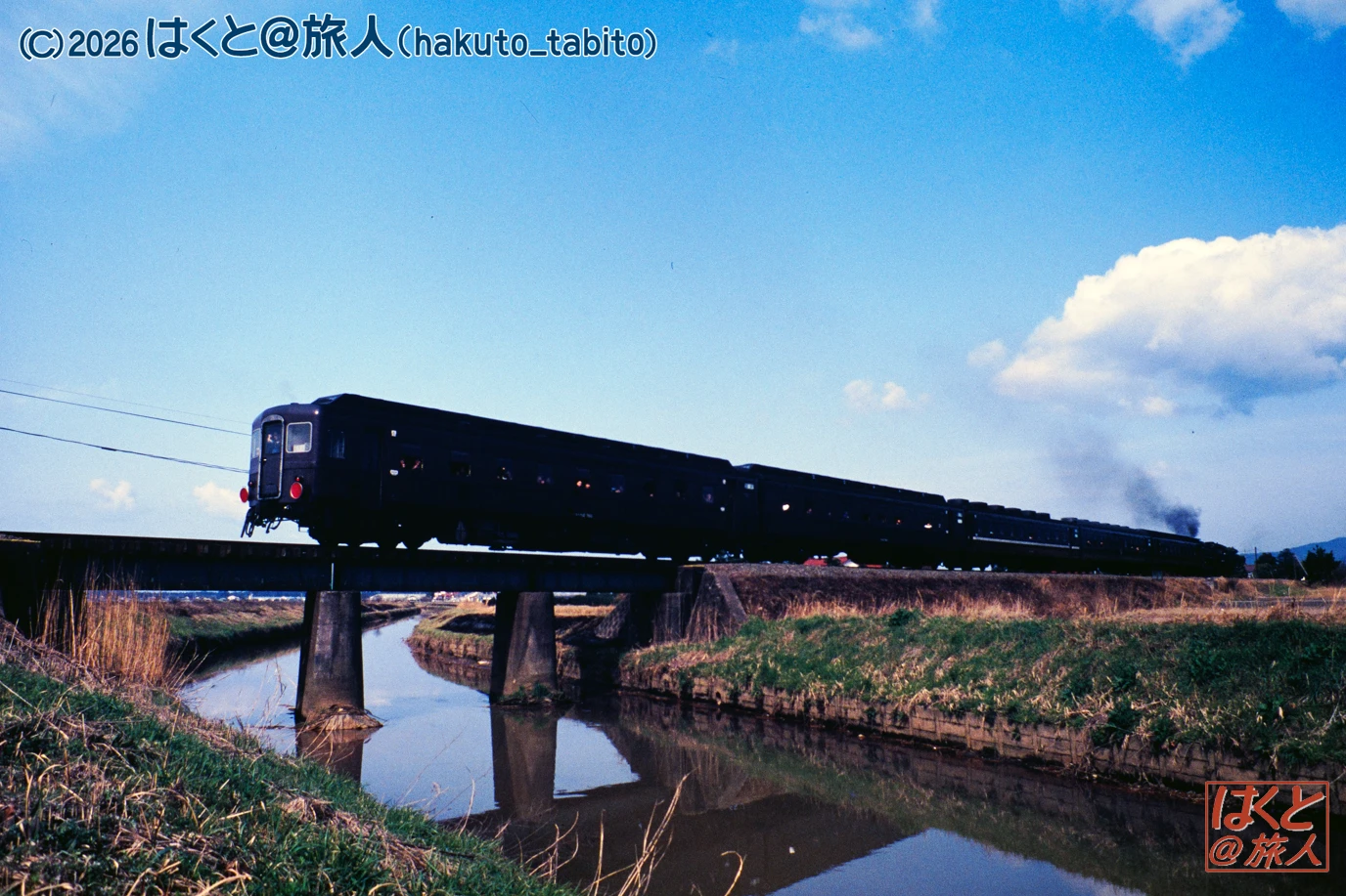 Black steam locomotive and passenger cars cross a bridge over a narrow canal in a rural field under a bright blue sky.