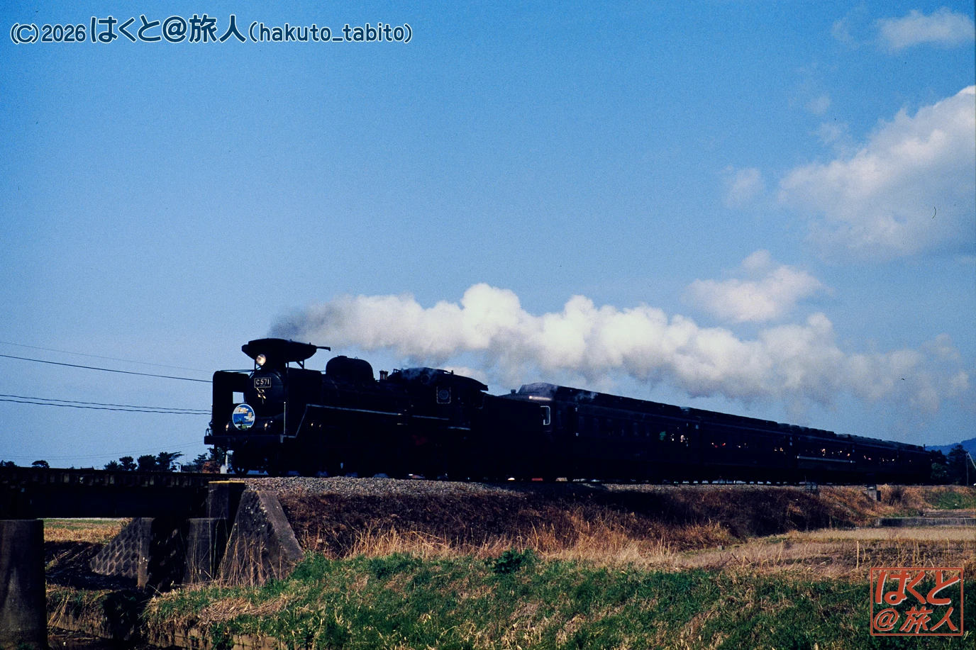 Black steam locomotive pulling a long train along a rural track under a blue sky with white clouds.
