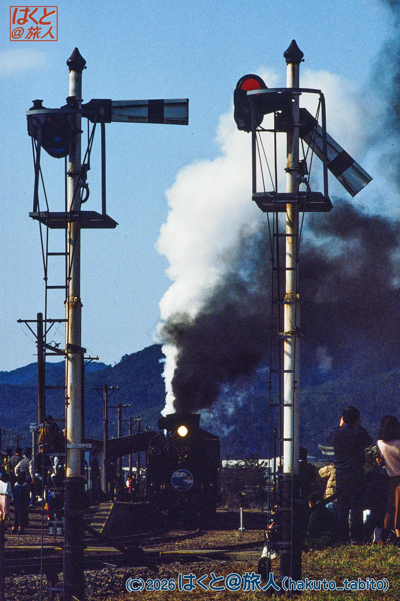 Steam locomotive at a rural train station with crossing signals and onlookers, mountains in the background.