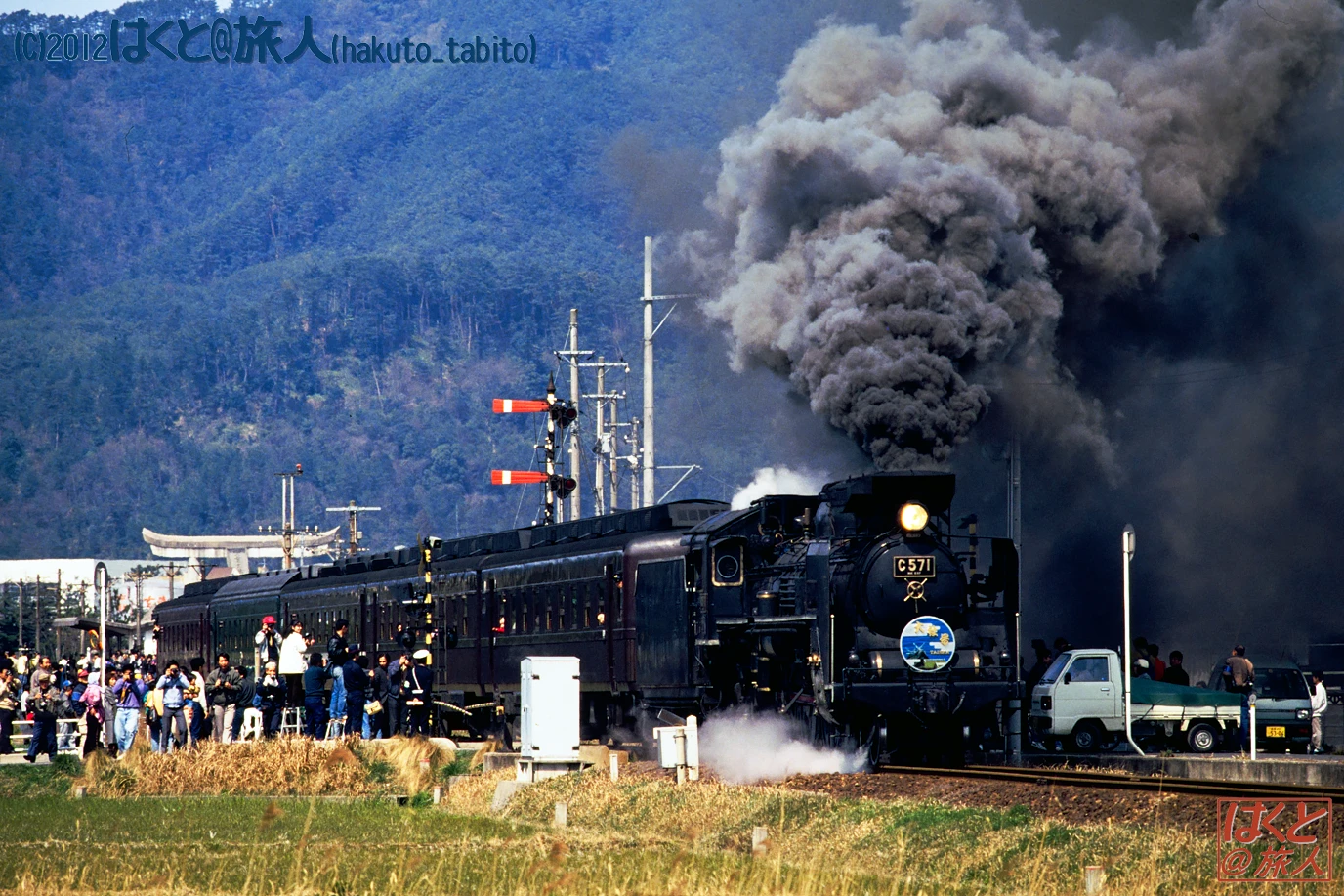 Steam locomotive C57 71 pulls into a rural station as a dense plume of smoke rises, with a crowd of onlookers along the tracks and a mountainous backdrop.
