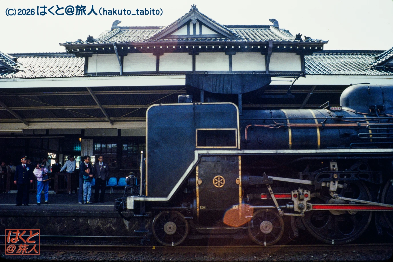 Steam locomotive at a station platform with onlookers, a traditional Japanese station building in the background.
