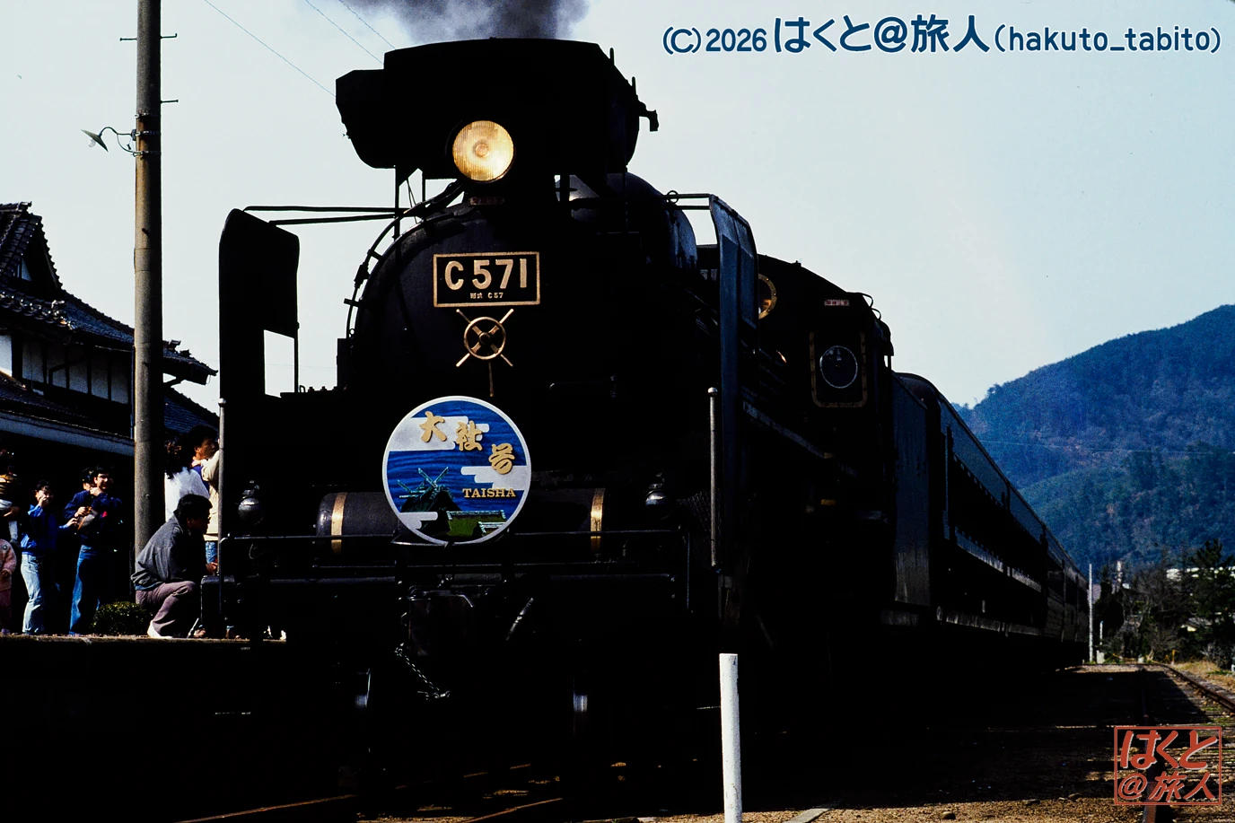 Steam locomotive C571 at a station, with bystanders on the left and mountains in the background.
