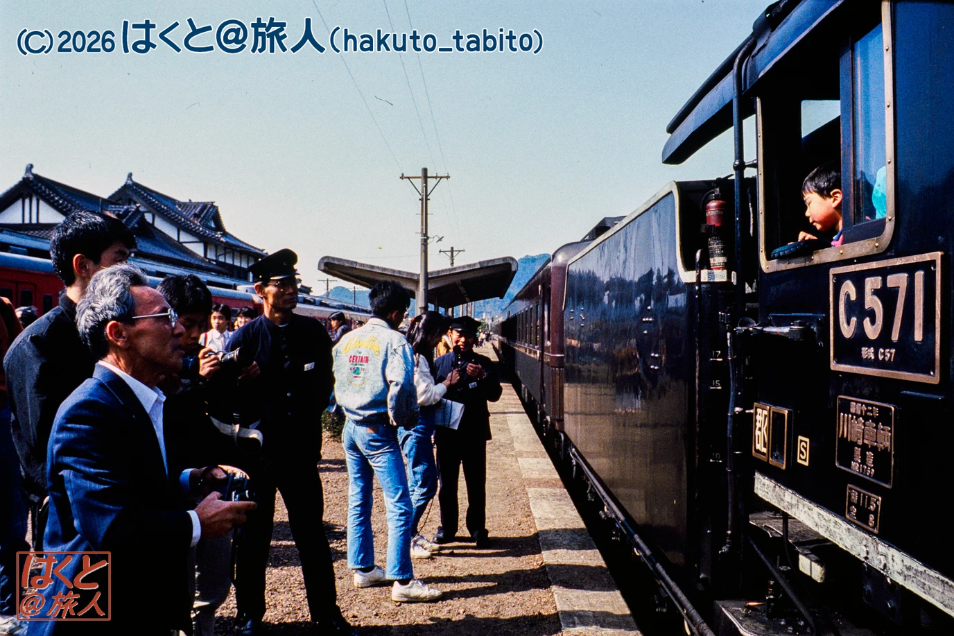 Crowd of people on a train platform beside a dark blue steam locomotive marked C571, preparing to depart at a sunny station.