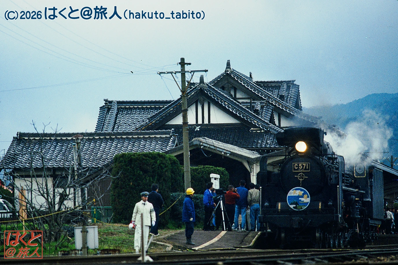 Steam locomotive C57 with steam rising, halting at a rural station beside a traditional tiled-roof Japanese building, with onlookers nearby.