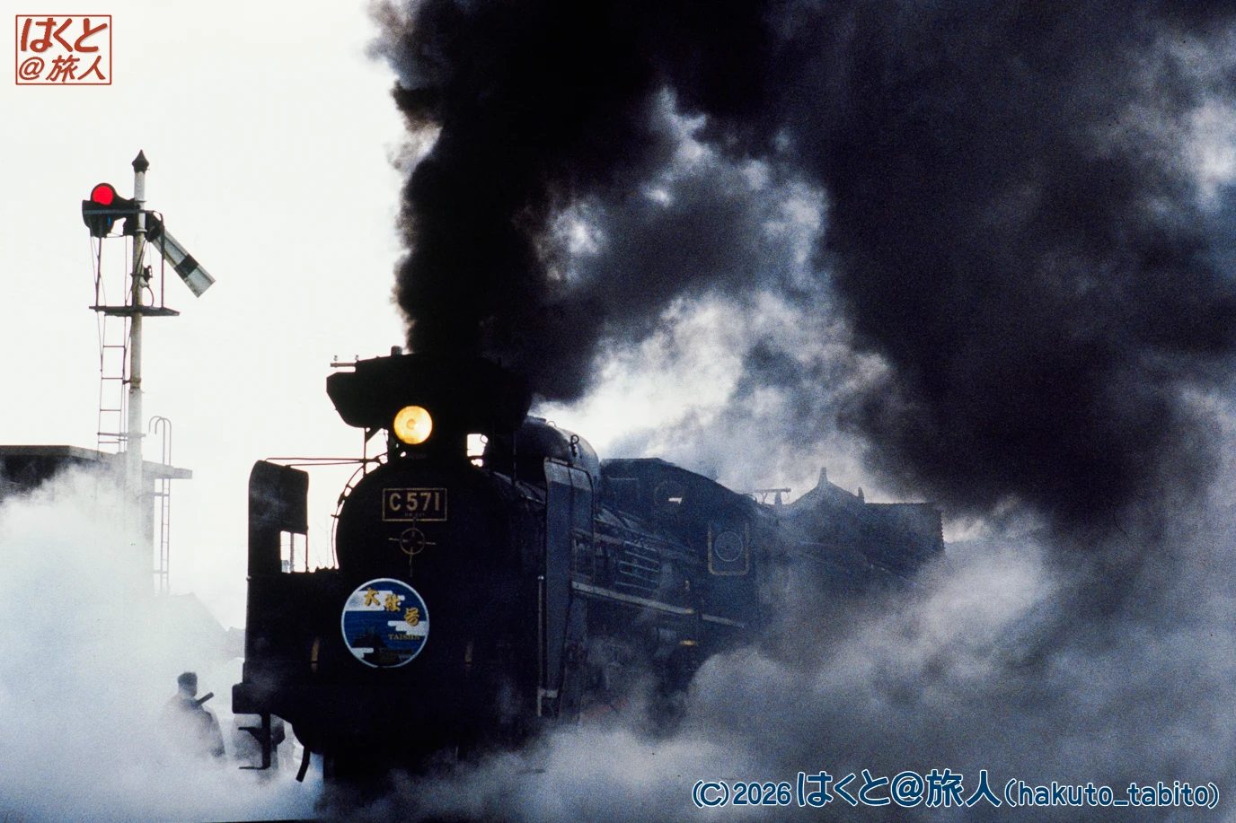 Steam locomotive C571 blasting through a foggy station with thick black smoke and a red signal nearby