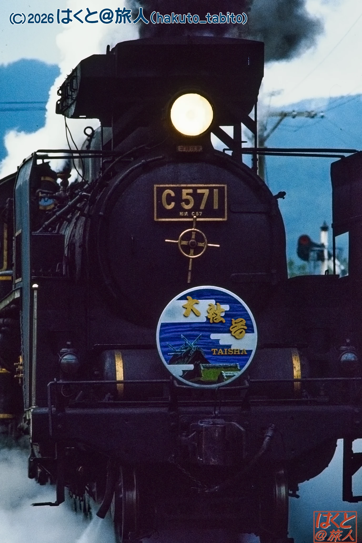 Front-on view of a black steam locomotive with a bright headlamp, bearing the number C571 and a round Taisha badge on the front plate.