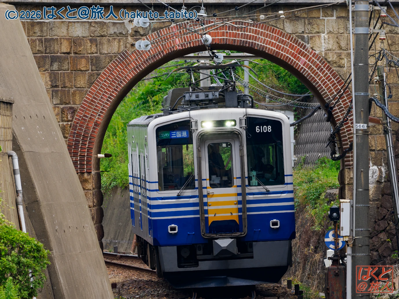 Commuter train in blue and white livery traveling under a brick arch tunnel, with overhead power lines above.