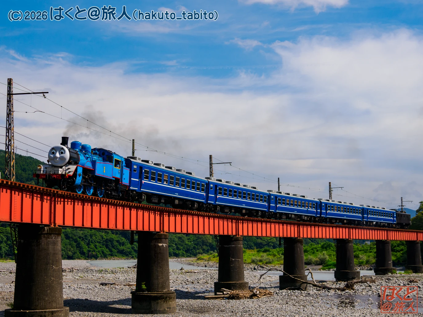 Blue steam locomotive pulling blue passenger cars crosses an orange steel bridge over a rocky riverbed with a bright blue sky behind it