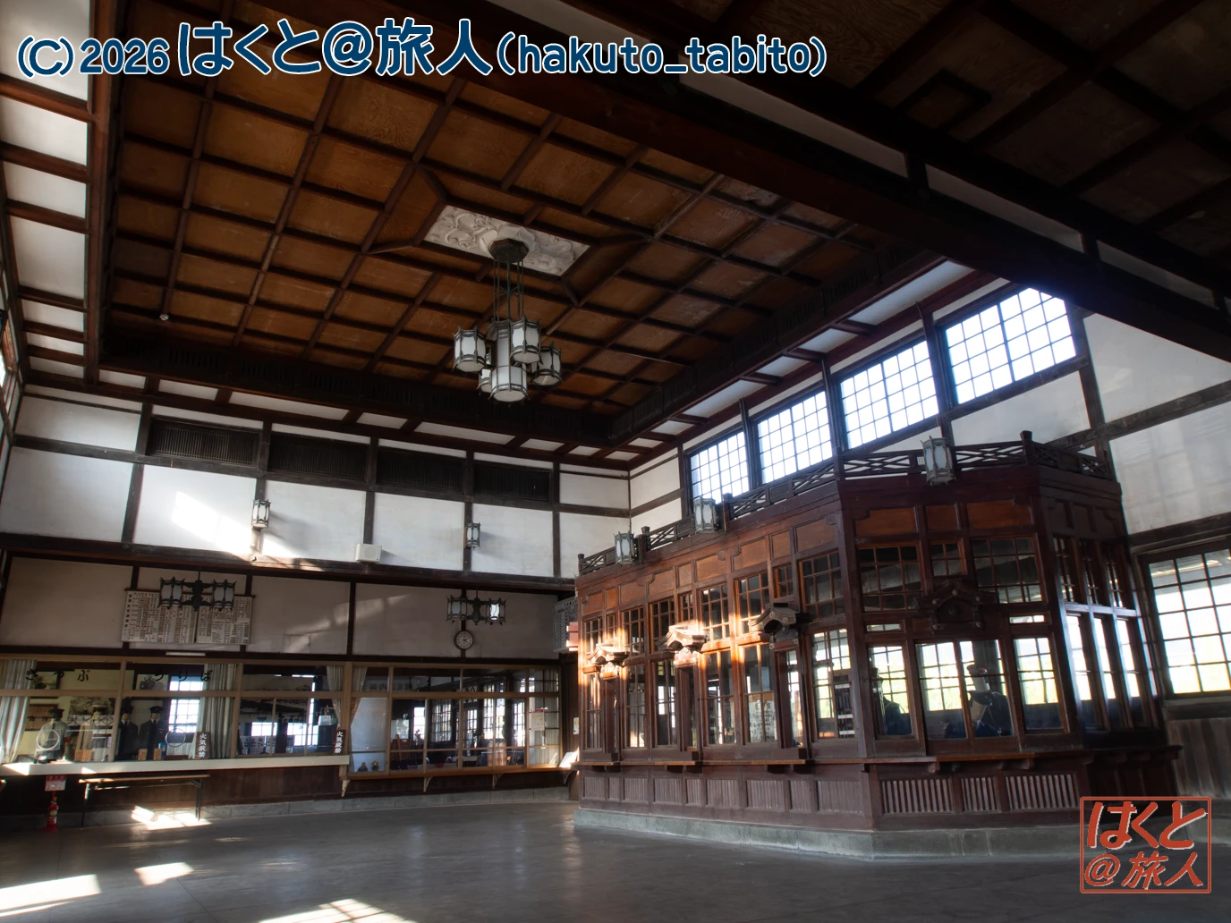 Expansive traditional Japanese interior with dark wooden beams, a large raised wooden display area on the right, grid windows along the upper walls, and hanging chandeliers; daylight streams in from the windows.