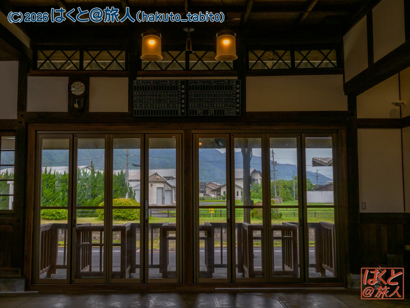 Interior of a traditional wooden room with tall glass doors opening to a rural street and distant mountains.