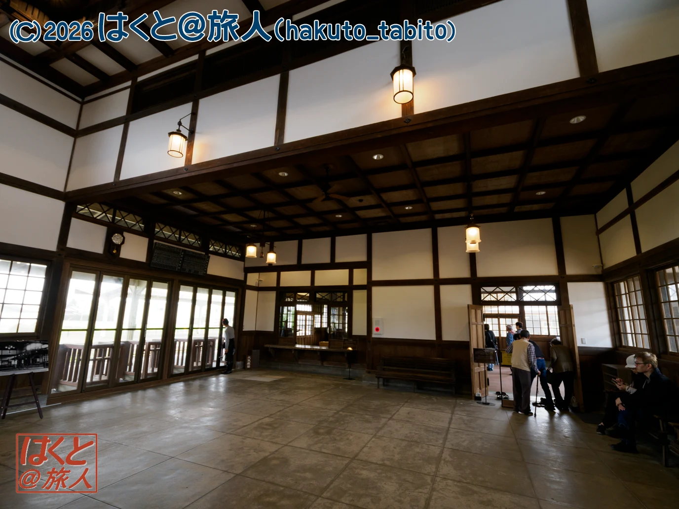 Spacious interior of a traditional Japanese-style hall with exposed dark wood beams, hanging lanterns, and people near the entrance.