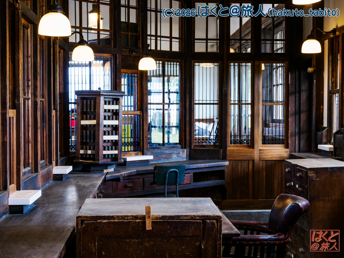 Interior of a vintage, wood-paneled room with desks, a green chair, and warm pendant lights seen through glass-paned windows