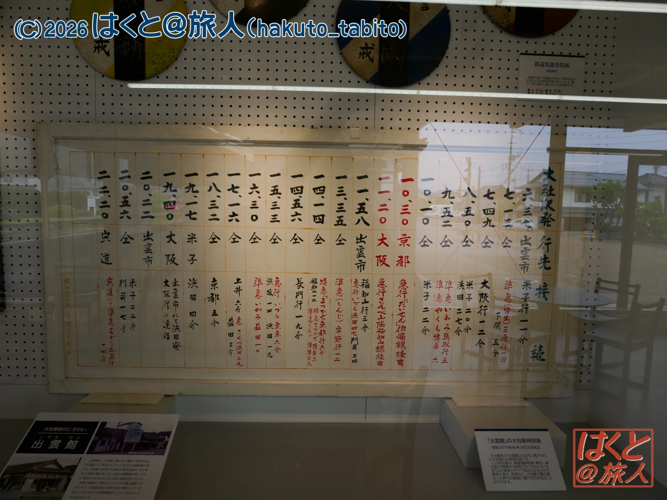 Framed Japanese timetable-like board with vertical columns of kanji characters, some in red, displayed in a glass case; reflections of a building visible.