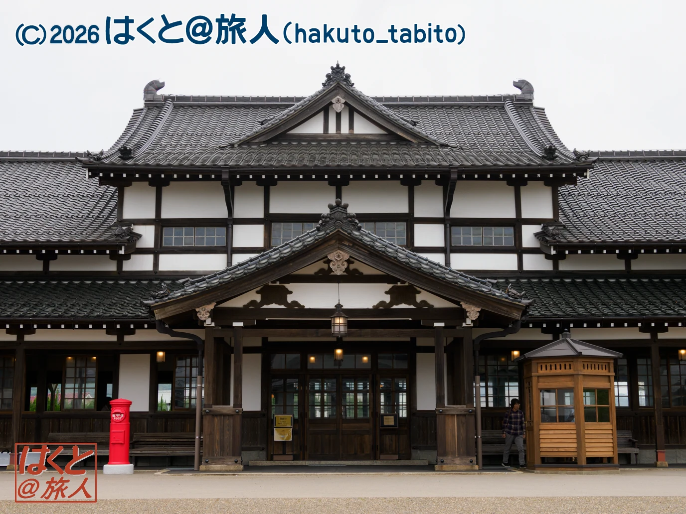 Front view of a traditional Japanese temple-style building with dark wooden beams, white plaster walls, and layered tiled roofs; a red mailbox sits at the left foreground and a wooden kiosk is on the right edge.